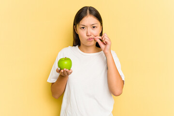 Young Asian woman eating an apple isolated on yellow background with fingers on lips keeping a secret.