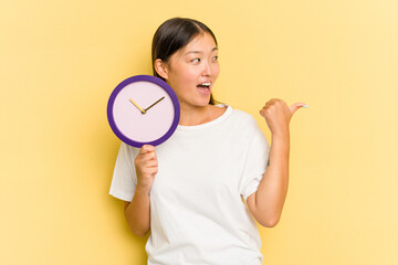 Young asian woman holding a clock isolated on yellow background points with thumb finger away, laughing and carefree.