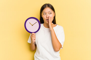 Young asian woman holding a clock isolated on yellow background is saying a secret hot braking news and looking aside