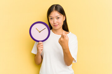 Young asian woman holding a clock isolated on yellow background pointing with finger at you as if inviting come closer.