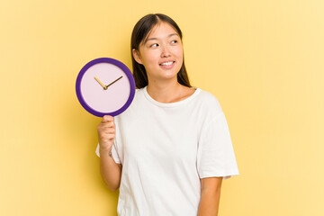 Young asian woman holding a clock isolated on yellow background looks aside smiling, cheerful and pleasant.