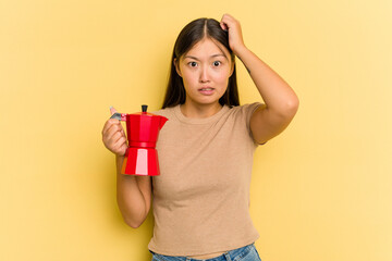 Young Asian woman holding coffee maker isolated on yellow background being shocked, she has...
