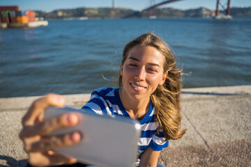 Positive woman taking selfie at the mobile phone while sitting at the embankment
