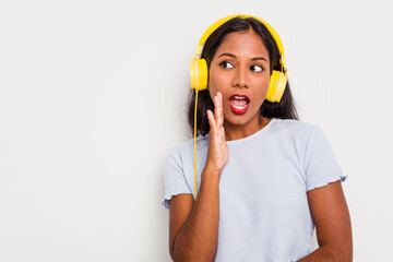 Young Indian woman listening to music wearing headphones isolated on white background is saying a secret hot braking news and looking aside