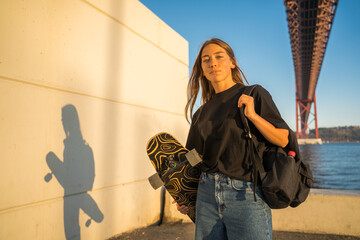 Confident woman carrying skateboard while posing at the embankment near the sea