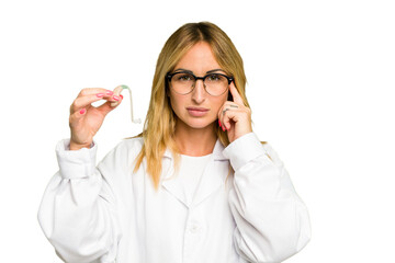 Young otorhinolaryngologist caucasian woman holding hearing aid isolated on green chroma background pointing temple with finger, thinking, focused on a task.