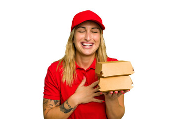 Young caucasian delivery woman holding a burger isolated on green chroma background laughs out loudly keeping hand on chest.