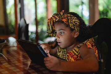 Imagen horizontal de una pequeña niña afro caribeña utilizando una tableta en el interior de su...