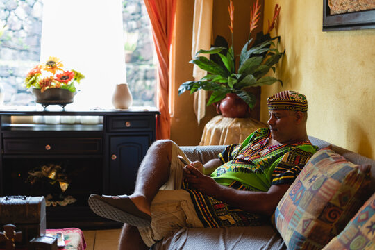 Imagen horizontal de un hombre latino adulto sentado cómodamente en el sofá de su sala utilizando su celular. 