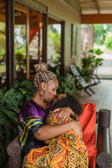 Imagen vertical de una madre afrocaribeña sentada cargando a su hija con mucho amor mientras la...