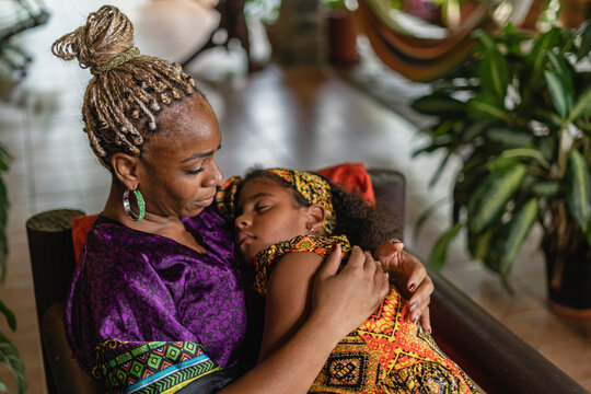 Imagen horizontal de una madre afrocaribeña sentada cargando a su hija con mucho amor mientras duerme. 