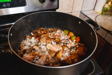 Imagen horizontal de una olla sobre la estufa llena de trozos de pollo junto a otros ingredientes en proceso de cocción al estilo  caribeño. 