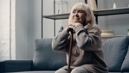 Fototapeta premium sad senior woman with dementia sitting on sofa while leaning on walking cane.