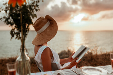 a young woman in a hat sits by the sea at sunset, reads a book and drinks champagne