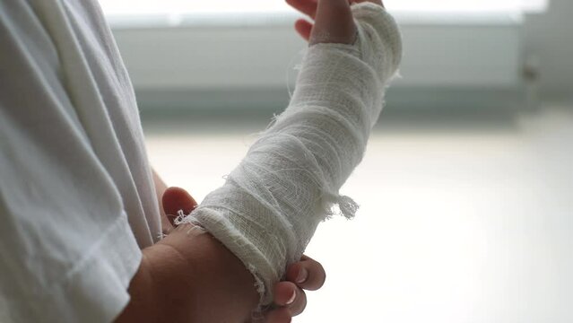 Side view of close-up cropped shot of unrecognizable little girl with broken hand wrapped in white plaster bandage gently massaging injured wrist on white background. Shooting in slow motion.