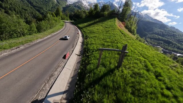 Aerial view spring sunny alpine forest resort infrastructure white sport car riding on slope serpentine road. FPV drone asphalt highway level automobile fast driving up mountain peak nature scenery 4k