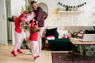 Merry Christmas concept. African American family celebrating the Christmas holiday, New Year. Daughters in Xmas pajamas meeting father with gifts at home.
