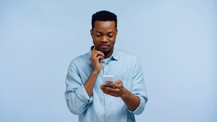 pensive african american man in shirt using mobile phone isolated on blue.