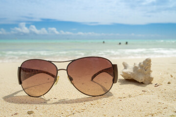 Fototapeta na wymiar Sunglasses lie on the beach next to a blurred sea shell. Dark glasses on the background of a blurred sea and a cloudy sky. Vacation concept.