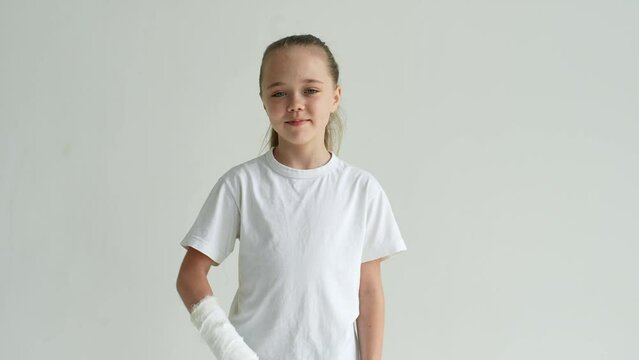 Studio portrait of positive smiling little girl with broken hand wrapped in white plaster bandage greeting waving hand looking at camera, standing on white isolated background. Shooting in slow motion