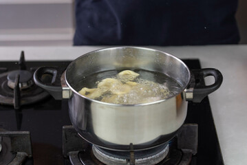 Chef cook hand making handmade tortellini ravioli with boiling water in pot saucepan on stove fire.
