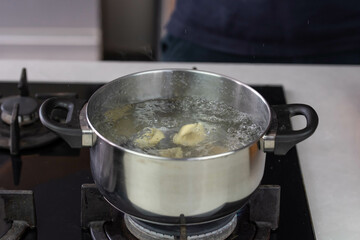 Chef cook hand making handmade tortellini ravioli with boiling water in pot saucepan on stove fire.
