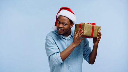 curious african american man in santa hat shaking christmas gift box isolated on blue.