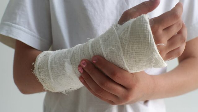 Close-up cropped shot of unrecognizable little girl with broken hand wrapped in white plaster bandage gently massaging injured forearm standing on white isolated background. Shooting in slow motion.