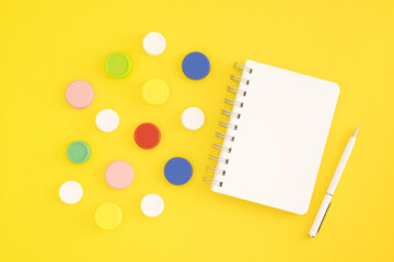 Top view of white notepad and colorful plastic bottle caps on yellow background