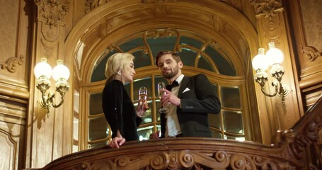 Two loving people dressed in elegant evening clothes enjoying cold champagne during conversation at casino place. Couple of celebrities standing on balcony with vintage interior around.