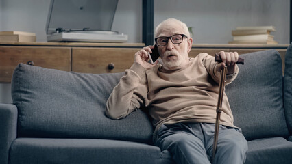 surprised senior man talking on smartphone and sitting on sofa with walking cane.