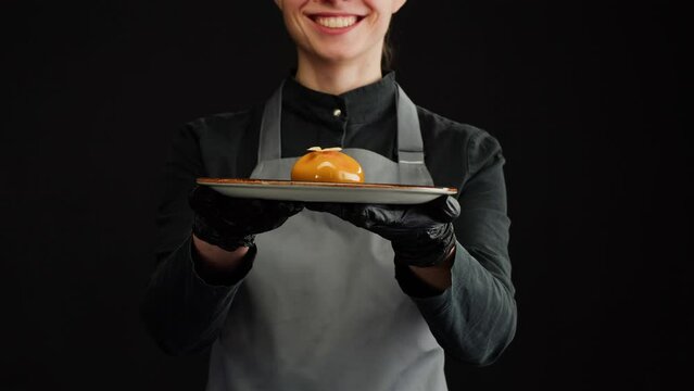Woman chef holding mousse orange cake, creamy dessert close-up. Almond dacquoise, raspberry confit, crispy layer with caramelized hazelnuts and raspberry powder, mousse based on bitter chocolate.