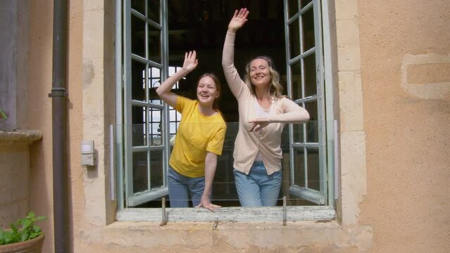 Cheerful woman and her daughter waving to somebody while looking from the window