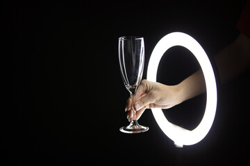 Female hand holding empty glass of champagne through led ring lamp on black background. Party concept