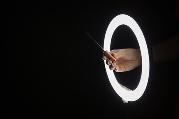 Female hands holding mascara brush through led ring lamp on black background. Beauty concept