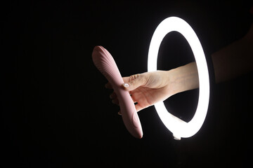 Female hand holding vibrator  through led ring lamp on black background. Sex concept - obrazy, fototapety, plakaty