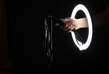 Female hand holding sex shop leather whip through led ring lamp on black background. Sex concept