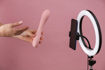 Sex blog. Female hands holding vibrator and broadcasting to smartphone camera with led rinng lamp on pink background
