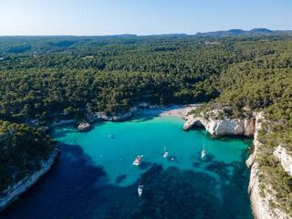 Fototapeta na wymiar drone views of virgin beach surrounded by vegetation, with people and boats moored, enjoying a day at the beach on the coast of Menorca.