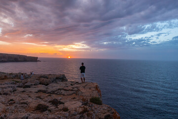 drone view with person standing on his back, watching the sunset on cliff, accompanied by more people on the coast of menorca.
