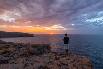 person standing on his back, watching the sunset on cliff, accompanied by more people on the coast of menorca.