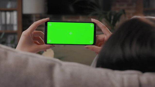 Over shoulder view of young woman holding smartphone with green mock-up screen horizontal mode. Woman lying on couch at home in evening, watching content videos blogs, news or films.Cinematic lighting