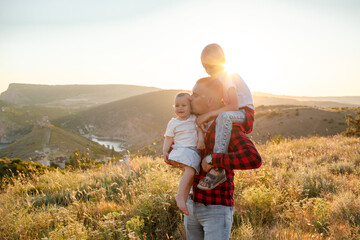 Happy family spending time together in the nature, father holding son on shoulders and daughter, have fun, playing with kid in vacation against the background the grass, mountain and sunset