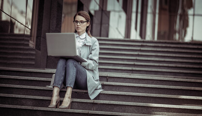 Fototapeta na wymiar Cute Caucasian woman is using laptop while sitting on stairs outdoors. Distant work. Modern business woman. Uses gadgets. Lifestyle