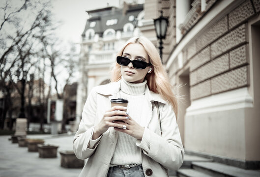 Young blonde woman in sunglasses and trench coat holding a cup of coffee on the go