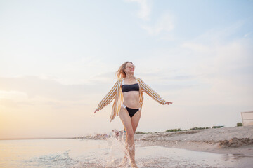 A beautiful girl is resting on the seashore against the background of sunset? the girl is wearing a swimsuit, sunglasses and a shirt