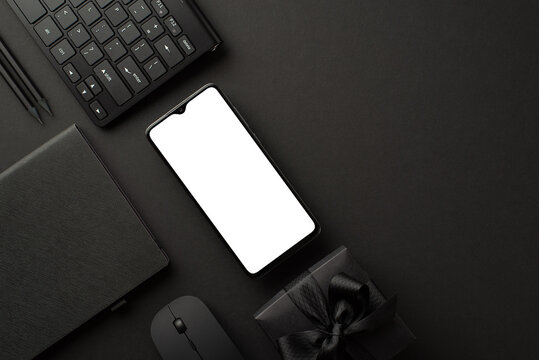 Black friday concept. Top view photo of black gift box with ribbon bow smartphone notepad pencils computer mouse and keyboard on isolated black background with blank space