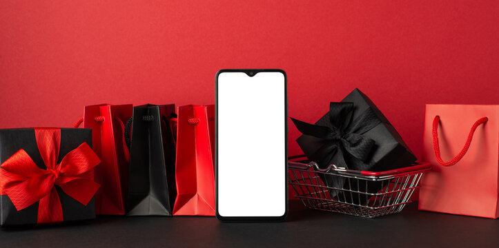 Black friday sales concept. Photo of smartphone red and black paper bags and giftbox with ribbon bow in shopping cart red wall background with empty space