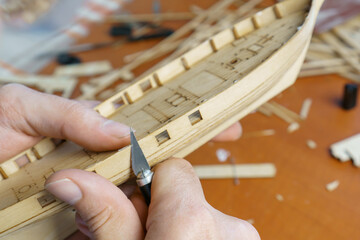 Hands of man cutting out details for ship model with clerical knife from plywood. Process of building toy ship, hobby and handicraft. Table with various materials, parts and devices for work - obrazy, fototapety, plakaty
