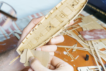 Hands of man adjusts plywood details for ship model, grinding on sandpaper. Process of building toy ship, hobby and handicraft. Table with various materials, parts and devices for work